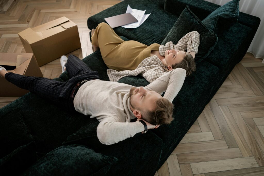 Expectant couple relaxing on a green sofa amidst moving boxes in their new home.
